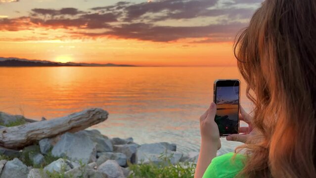 a teenage girl in a Green T-shirt stands with her back to the camera and shoots a beautiful sunset on Pacific Ocean she holds an iPhone 13 phone in her hands blond red hair stones setting sun sea sky