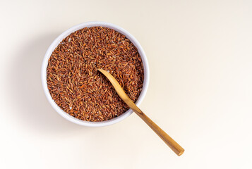 Uncooked red rice in a ceramic bowl with wooden spoon isolated on white background, top view