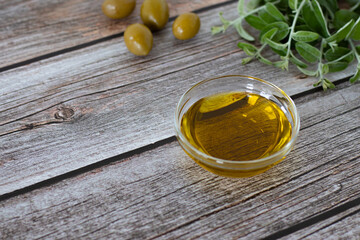 Natural cooking greek olive oil in a glass bowl with green olives and fresh tree branches on a wooden background with copy space. A close-up.