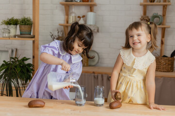 two little girls in the kitchen pour milk from a glass jug into glasses and drink. space for text © Tina kids photo