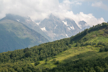 Panoramic view of the green meadow with incredible snowy mountains crossed by big white clouds, in the Mestia-Ushguli trek, Georgia.