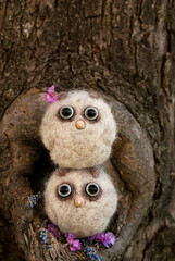 Two small owlets made of wool with beady eyes and a beak made of plastic in light colors sit near a small hollow in a tree with flowers
