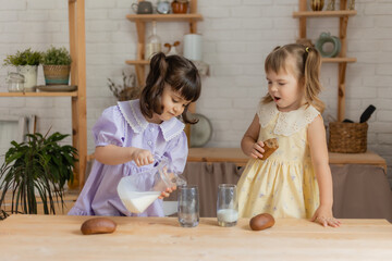 two little girls in the kitchen pour milk from a glass jug into glasses and drink. space for text