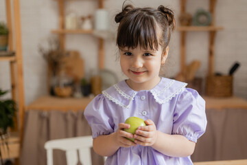 Fototapeta premium charming little girl in a lilac dress eats a green apple in the kitchen. space for text, banner