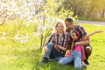 Outdoor portrait of happy young family playing in spring park under blooming tree, lovely couple with little child having fun in sunny garden