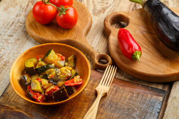 A plate of grilled vegetables next to tomatoes and eggplant and peppers on wooden boards next to a wooden fork.