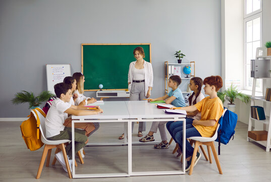Children having class in cozy classroom interior with green chalkboard. Happy students sitting around big table, reading books, writing in notebooks and talking with teacher. Back to school concept
