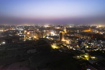 aerial drone dusk twilight shot showing orange lights of streets, homes and markets surrounding a skyscraper with the city scape stretching into the distance in gurgaon haryana delhi