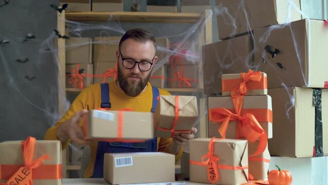 young bearded man in glasses business owner sits in halloween decorated home office. Folds boxes with orange bows. concept of selling goods for the holiday of all the dead, discounts and promotions.