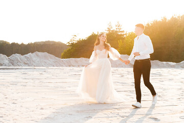 Fototapeta na wymiar Amazing newlyweds in love, holding hands, run along a sandy quarry at sunset.