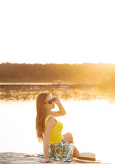 A stylish brunette in bright summer clothes is resting on the shore of the lake at sunset