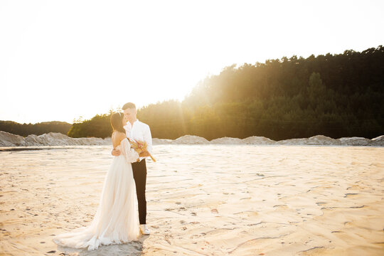 Luxury bride in a white dress ivory shade. Sweet couple is embracing tenderly against the background of white sand at sunset