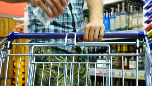 Close-up of a shopping trolley in strong drinks department and a man puts a bottle of vodka into it