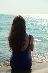a beautiful, slender woman with curly hair is sitting on the beach on the sand in sunny weather, sunbathing and enjoying her vacation with sunglasses on her face looking towards the sea