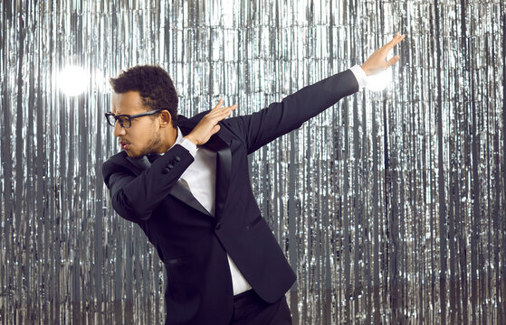 Happy Man In An Elegant Dinner Suit Having Fun At A Modern Fashion Party. Confident Energetic Young Black Guy In A Tuxedo Outfit And Glasses Dancing Against A Silver Foil Fringe Background