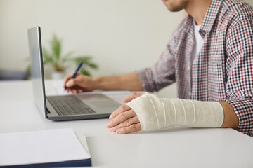 Young student or business man with elastic bandage wrapped around his hand sitting at desk, working on laptop computer, studying new information and taking notes. Cropped shot. Wrist injury concept