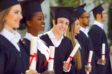 Young smiling man graduate participates in graduation ceremony and looks at camera with smile dressed in graduation gown standing among multiracial college students outdoors. Selective focus