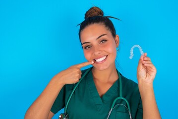 beautiful doctor woman wearing medical uniform over blue background holding an invisible aligner and pointing perfect straight teeth. Dental healthcare and confidence concept.