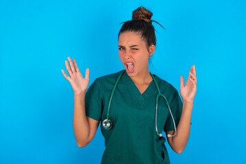 Crazy outraged beautiful doctor woman wearing medical uniform over blue background screams loudly...