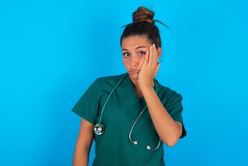 Sad lonely beautiful doctor woman wearing medical uniform over blue background touches cheek with...