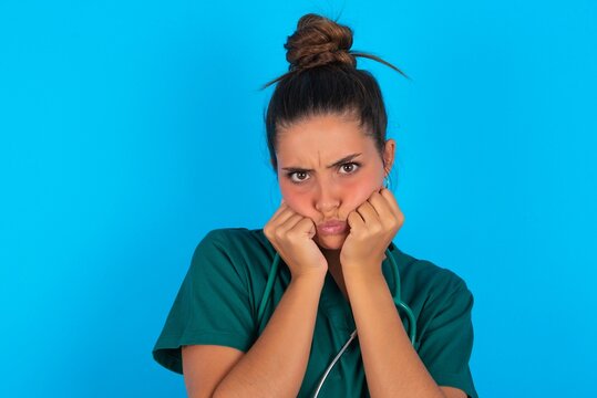 beautiful doctor woman wearing medical uniform over blue background with surprised expression keeps hands under chin keeps lips folded makes funny grimace