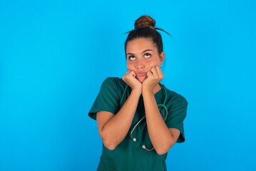 Portrait of sad beautiful doctor woman wearing medical uniform over blue background hands face look empty space