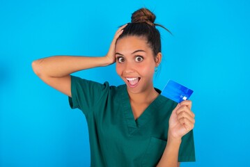 Impressed funny beautiful doctor woman wearing medical uniform over blue background hand on head holding bank card
