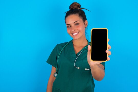 Charming adorable beautiful doctor woman wearing medical uniform over blue background holding modern device, showing black screen smartphone