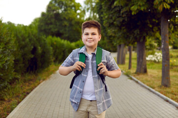 Cheerful Caucasian boy teenager posing with school backpack on back walks after study day symbolizes happy childhood dressed in casual clothes, stands in summer green park. Selective focus