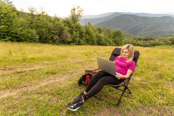 Naklejka na ściany i meble A woman in a sports warm suit works on a laptop outdoors in a mountainous area.