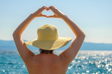 Lady in straw hat stands against blue sea showing heart shaped hands over head. Young woman looks...