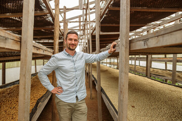 Happy male manager working at coffee farm and standing near wooden structure for drying coffee beans. Honey dry process