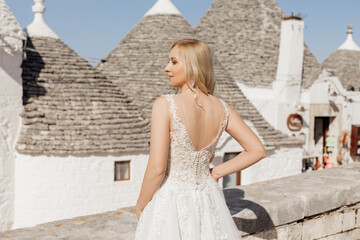 Back view of young happy woman bride standing near conical roofs of trullo in sunny Italy, putting...