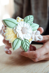 Hand of woman holding and showing closeup beautiful icing flower shaped cookie on table in cafe, blurred background. Catering, coffee house, serving food, dessert, sweet gingerbread