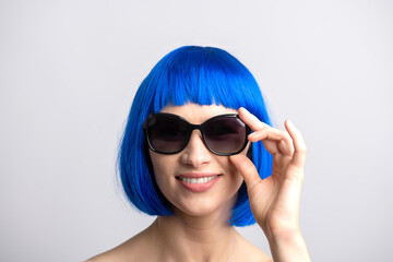 Beauty, fashion concept. Portrait of beautiful and sexy woman with blue wig and sunglasses looking to camera with smile. Sun tan soft skin, pink lipstick and white teeth. Model with naked shoulders