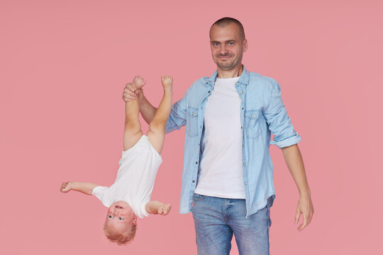 Young father holding little son by the leg upside down on pink background