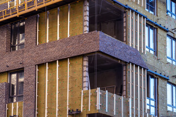 Monolithic concrete frame of apartment building under construction with partially attached facade cladding blocks. Energy efficiency. Installation of external wall thermal insulation with rock wool.
