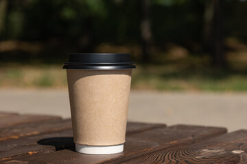 A paper cup of coffee with a lid stands on a rustic bench table against the backdrop of green foliage and sandy beach, sunny morning, breakfast, coffee break, coffee to go - blurred bokeh background