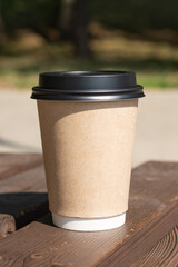 A paper cup of coffee with a lid stands on a rustic bench table against the backdrop of green foliage and sandy beach, sunny morning, breakfast, coffee break, coffee to go - blurred bokeh background