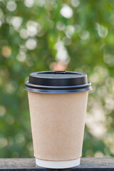 A paper siakanchik of coffee with a lid stands on a metal railing against the background of green foliage, sunny morning, breakfast, coffee break, coffee to go - blurred bokeh background, vertical