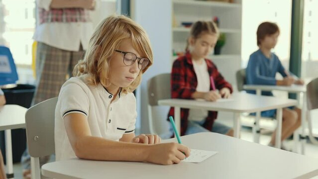 Red-head pupil wearing glasses sits at a desk at school and fills out tests, and the teacher in the background gives an assignment. People and educational concept.