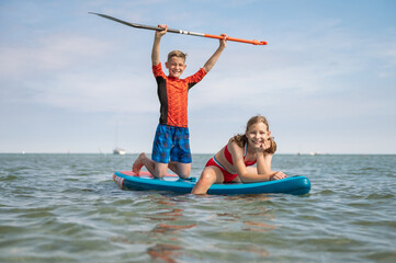 Happy teenage siblings having fun on paddle board at sunny summer day at vacation on sea