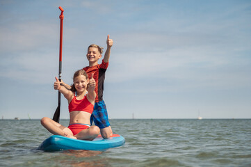 Happy teenage siblings having fun on paddle board at sunny summer day at vacation on sea