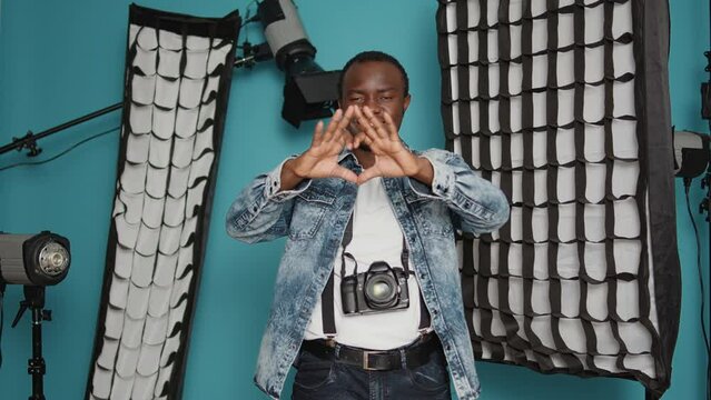 African american photographer doing heart shape symbol with hands, psoing with romantic love gesture over background with photography lighting equipment. Valentines day feelings sign.