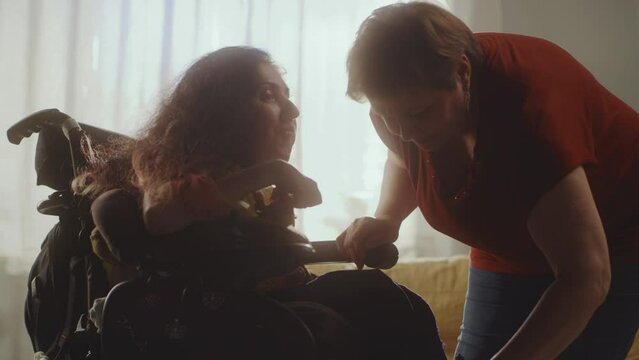Aged woman caregiver helping woman with a spinal muscular atrophy setting a modern motorized wheelchair for comfortable sitting