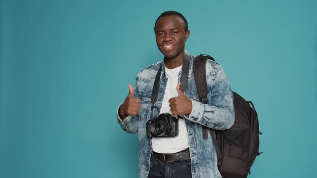 Positive photographer doing thumbs up gesture in studio, doing approval and agreement sign. Having backpack to leave on urban cityspace adventure and take pictures with dslr camera.