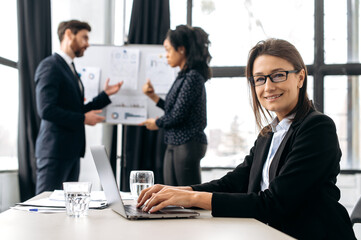 Confident elegant caucasian business woman with glasses, sitting at the desk at conference, near laptop. Beautiful elegant female employee on briefing meeting, looking at the camera, smiling friendly