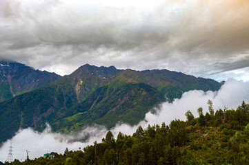 Fototapeta premium Fog in the mountains. Kalpa, Himachal Pradesh, India. Elevated view of Kalpa village dwarfed by the snow-capped Himalayas under a cloudy and overcast sky at dawn in Kalpa, Himachal Pradesh, India.