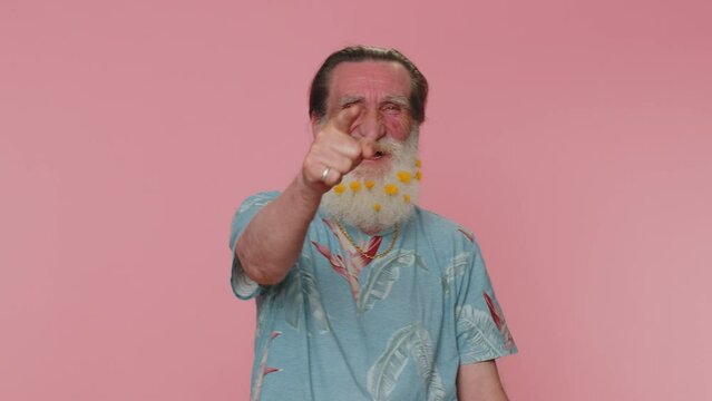 Amused senior man flowered beard pointing finger to camera, laughing out loud taunting making fun of ridiculous appearance, funny joke. Elderly grandfather posing alone on pink studio wall background