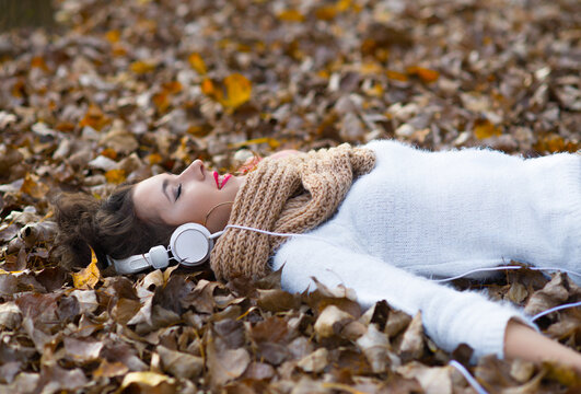 Young woman listening music through headphones while lying in autumn leaves